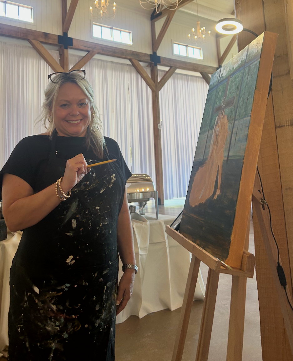 Samantha painting at an easel at a wedding venue, brush in hand, chandeliers and timber trusses behind her.